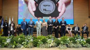 A imagem mostra o palco de um auditório do TJTO. Ao centro, sete autoridades, entre homens e mulheres, estão em pé, algumas segurando medalhas comemorativas. A segunda pessoa da esquerda para a direita é o desembargador Osmar Duarte Marcelino, vestido com terno cinza, camisa branca e gravata azul. Ao fundo, outras autoridades estão sentadas. Atrás, um grande telão exibe a imagem de mãos segurando uma medalha. À esquerda do palco, aparecem bandeiras. Na parte frontal, aparecem arranjos de plantas e flores nas cores verde, branco e azul.