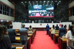 A imagem mostra o Plenário Juscelino Kubitschek da Assembleia Legislativa de Minas Gerais. Em primeiro plano, várias pessoas estão de pé entre as fileiras de mesas e cadeiras, voltadas para a mesa de autoridades. Ao fundo, autoridades estão alinhadas atrás de uma longa mesa branca também permanecem de pé. À esquerda da imagem aparece a bandeira do Brasil e, à direita, a bandeira de Minas Gerais. Acima, um grande telão exibe a transmissão ao vivo da mesma cena. O ambiente possui carpete vermelho no chão e paredes revestidas em mármore escuro