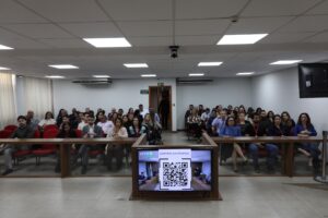 A imagem mostra os servidores do TJMMG durante reunião interna sobre as Metas Nacionais do Poder Judiciário para 2027. Eles estão sentados em cadeiras vermelhas do Plenário do TJMMG. As paredes ao fundo são brancas e há uma bancada de madeira em primeiro plano