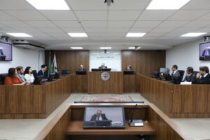 A imagem mostra desembargadores e juízes de Direito do Juízo Militar durante reunião interna sobre as Metas Nacionais do Poder Judiciário para 2027, sentados em uma tribuna de madeira. Eles estão posicionados atrás de microfones e computadores, todos vestidos com trajes sociais. O ambiente apresenta paredes revestidas em madeira e bandeiras oficiais ao fundo.