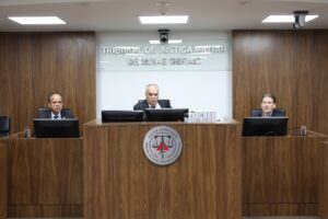 A imagem mostra os desembargadores no Plenário do TJMMG, durante reunião interna sobre as Metas Nacionais do Poder Judiciário para 2027, sentados em uma tribuna de madeira. Eles estão posicionados atrás de microfones e computadores, todos vestidos com trajes sociais. O ambiente apresenta paredes revestidas em madeira.