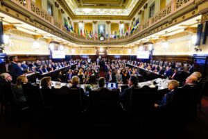 A imagem mostra o Salão Nobre da sede TJSP repleto de autoridades convidadas para a abertura do XX Encontro do Consepre. Um grupo de autoridades está disposto em semicírculo, enquanto outro está sentado ao centro. O salão tem paredes com pé direito alto, com um mezanino, tudo em tom claro com detalhes dourados e madeira. No mezanino estão dispostas bandeiras de todos os estados com representatividade no Consepre