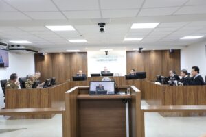 A imagem mostra os desembargadores durante a Sessão de Julgamento no Tribunal Pleno, sentados em uma tribuna de madeira. Eles estão posicionados atrás de microfones e computadores, alguns com trajes militares, outros de toga e um de camisa social e gravata. O ambiente apresenta paredes revestidas em madeira, bandeiras oficiais e o brasão do TJMMG ao fundo.
