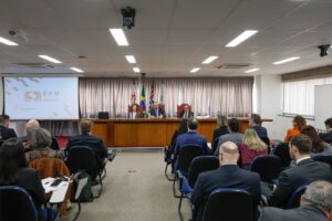 A imagem mostra uma sala institucional do TJSP. Em primeiro plano, aparecem diversas pessoas sentadas em fileiras de cadeiras, de frente para uma mesa de autoridades. Ao fundo, três pessoas estão sentadas à mesa, diante de bandeiras e tendo à esquerda da imagem uma tela de projeção.
