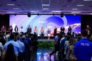 A imagem mostra o palco da solenidade de abertura do 6º FestLabs nacional. Um grupo de pessoas está reunido diante de um grande telão, que exibe a identidade visual do evento, nas cores azul e roxo. Nele está escrito “sexto Fest Lebs”. Em primeiro plano, aparece uma plateia voltada para o palco.