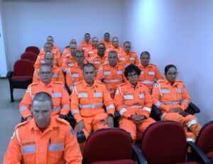 A imagem mostra os alunos da Academia de Bombeiros Militar durante sessão de julgamento do Tribunal Pleno. Todos vestem uniformes do bombeiro militar na cor laranja. Eles estão sentados em cadeiras vermelhas do Plenário do TJMMG. As paredes ao fundo são brancas.