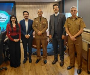 A imagem mostra cinco pessoas, na Sala Diamante do TJMMG, em pé, lado a lado. Da direita para a esquerda, está o tenente-coronel Jean Carlo Alcântara Pedra, com uniforme da PMMG marrom; o desembargador Fernando José Armando Ribeiro, vestindo terno cinza; o coronel Halysson Claudino Câmara dos Santos, com uniforme da PMMG marrom; o servidor Lucas Figueiredo de Oliveira, usando terno azul-marinho e camisa branca; e a servidora Ana Flávia Taeko Reis Miyashita, usando óculos, blusa vermelha e calça preta. O ambiente possui paredes e pisos revestidos em madeira. 