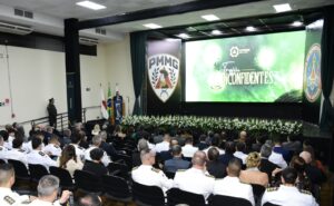 A imagem mostra o interior do auditório do Clube dos Oficiais da Polícia Militar e do Corpo de Bombeiros Militar de Minas Gerais. Em primeiro plano, há diversas pessoas sentadas em fileiras de cadeiras, entre elas militares trajando uniformes brancos. Ao fundo, aparece um telão com a identidade visual do evento, escrito “AOPMBM, Minas Gerais, Troféu Inconfidentes”. Nas laterais do telão, aparecem grandes painéis com os brasões da PMMG e do CBMMG. No palco, um homem está em pé diante de uma tribuna, tendo ao fundo três bandeiras. A decoração do palco é formada por arranjos de flores brancas.
