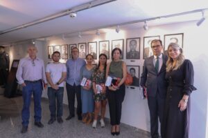 A imagem mostra um grupo de pessoas, entre homens e mulheres, em pé no Memorial da Justiça Militar. Ao fundo, aparece uma parede com diversas reproduções de retratos em preto e branco de integrantes da Justiça Militar, tendo em destaque o desembargador Laurentino de Andrade Filocre.