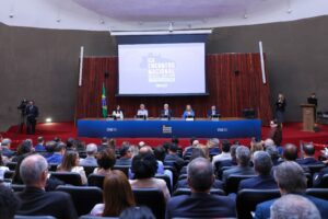 A imagem mostra um auditório do Tribunal Superior Eleitoral. Em primeiro plano, aparece o público sentado em fileiras de cadeiras. Ao fundo, no palco, cinco pessoas estão sentadas atrás de uma mesa, coberta por um painel azul com a identidade visual do evento. Ao centro, aparece uma tela de projeção escrito 20º Encontro Nacional do Poder Judiciário – 2ª Reunião Preparatória”.