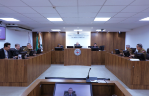 A imagem mostra os desembargadores durante a Sessão de Julgamento no Tribunal Pleno, sentados em uma tribuna de madeira. Eles estão posicionados atrás de microfones e computadores, alguns com trajes militares, outros de toga. O ambiente apresenta paredes revestidas em madeira, bandeiras oficiais e o brasão do TJMMG ao fundo.