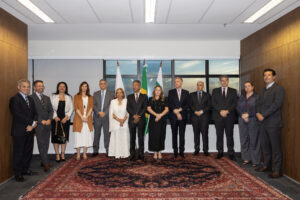 A imagem mostra a composição do dispositivo de honra durante a cerimônia de posse das novas ouvidoras do TJMG, no Gabinete da Presidência, um ambiente formal com paredes laterais revestidas de madeira, piso claro com um grande tapete a avermelhado ao centro e janelas ao fundo, tendo à frente três bandeiras hasteadas. O grupo é formado por 13 autoridades dispostas em pé, lado a lado, sendo homens e mulheres que vestem trajes formais. A ouvidora, desembargadora Maria Luiza Santana Assunção, é a sexta pessoa na imagem, e veste um conjunto totalmente branco composto por blazer, blusa interna e saia longa rendada. A ouvidora-adjunta, desembargadora Ivone Campos Guilarducci Cerqueira, está na sequência, trajando um vestido preto de comprimento midi. O desembargador Fernando José Armando Ribeiro, ouvidor do TJMMG, é o primeiro homem à direita da imagem, e veste um terno completo cinza-escuro, camisa social clara e gravata. 