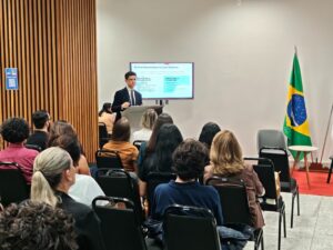A imagem mostra o juiz de Direito George Wálter Barreto Paviótti falando em uma tribuna, diante de um público sentado em fileiras de cadeiras, no TRF6. Ele veste terno e gravata escuros, e camisa branca. Ao fundo, há uma tela exibindo slides da apresentação. O ambiente possui uma parede clara e outra revestida em madeira, e a bandeira do Brasil está posicionada à direita da imagem.