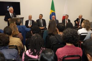 A imagem mostra, em primeiro plano, várias pessoas sentadas em cadeiras no TRF6. Ao fundo, aparecem cinco homens sentados em cadeiras, todos vestindo ternos e gravatas e voltados para o público. Entre eles, o primeiro é o desembargador Rúbio Paulino Coelho, que veste terno preto, camisa branca e gravata preta. Atrás dos homens, aparecem duas bandeiras. À esquerda da imagem, há um homem em pé, falando em uma tribuna.