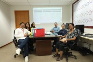 A imagem mostra cinco pessoas em uma sala do setor de gestão documental TJMMG, sentadas ao redor de uma mesa de reunião com pastas e papéis. Ao fundo, há uma projeção na parede.