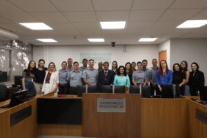 A imagem registra um grupo de pessoas, entre homens e mulheres, que integram a 4ª AJME. Todos estão em pé, posicionados atrás de uma bancada de madeira. Na parede atrás deles está uma placa de acrílico escrito: Quarta auditoria. Justiça Militar de Minas Gerais. Ao centro, está o juiz André de Mourão Motta, que veste terno preto, camisa branca e gravata listrada nas cores vermelho, cinza e preto. Na imagem, ao lado do juiz está a juíza Renata Rodrigues de Pádua, de camisa azul-claro. As demais pessoas da imagem são os representantes do CBMMG, vestindo uniformes militares da corporação, além de servidoras da Auditoria, vestidos formalmente.