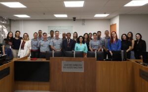 A imagem registra um grupo de pessoas, entre homens e mulheres, que integram a 4ª AJME. Todos estão em pé, posicionados atrás de uma bancada de madeira. Na parede atrás deles está uma placa de acrílico escrito: Quarta auditoria. Justiça Militar de Minas Gerais. Ao centro, está o juiz André de Mourão Motta, que veste terno preto, camisa branca e gravata listrada nas cores vermelho, cinza e preto. Na imagem, ao lado do juiz está a juíza Renata Rodrigues de Pádua, de camisa azul-claro. As demais pessoas da imagem são os representantes do CBMMG, vestindo uniformes militares da corporação, além de servidoras da Auditoria, vestidos formalmente.
