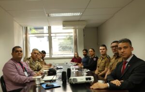 A imagem mostra um grupo de 10 pessoas sentadas em uma mesa de reunião do TJMMG, entre homens e mulheres vestidos com roupas formais e uniformes da PMMG. À direita, próximo à janela, está a juíza de Direito Carolina Aleixo Benetti de Oliveira Rodrigues, vestindo blusa vermelha.
