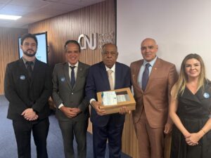 A imagem mostra cinco pessoas em pé, na sede do CNJ. A segunda pessoa, da esquerda para a direita, é o desembargador Jâmes Ferreira Santos, vestindo terno cinza, camisa branca e gravata azul. Ao seu lado, está o ministro Benedito Gonçalves, usando terno azul, camisa branca e gravata bege. Ao lado dele, está o desembargador Raimundo Messias Júnior, vestindo terno marrom, camisa azul-clara e gravata escura. À direita, está a servidora Tatiana Reis Teixeira, usando vestido preto. À esquerda, está o servidor Tiago Augusto Duarte Pereira, usando terno preto, camisa cinza e gravata preta. Ao fundo, aparece uma parede revestida em madeira e a logomarca do CNJ.