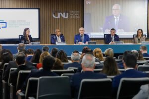 A imagem mostra a solenidade de encerramento do Encontro Nacional Integração de Boas Práticas: Diálogos entre o CNJ e o Colégio de Corregedores e Corregedoras da Justiça do Brasil, no auditório do CNJ, em Brasília. Em primeiro plano está a plateia sentada em poltronas escuras e de frente para a mesa de abertura. A mesa, azul, está em frente a um painel de madeira onde se destaca o logotipo em relevo cinza do CNJ. Entre as autoridades, há três homens e duas mulheres. O homem ao centro é o presidente do CNJ e do STF, ministro Edson Faquin, de terno e gravata azul, e óculos. Ao lado esquerdo dele na imagem está o corregedor nacional de Justiça, de terno azul e gravata clara.