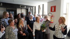 A imagem mostra um grupo de pessoas, entre homens e mulheres, reunidas em um espaço interno da Amagis durante a inauguração do Espaço Amor à Vida. À direita, a juíza Flávia Birchal conversa com os convidados e gesticula com as mãos. Ao lado dela, a juíza de Direito titular do Juízo Militar Carolina Aleixo Benetti de Oliveira Rodrigues está encostada em uma parede branca. Outras pessoas aparecem reunidas ao fundo, acompanhando o momento. 