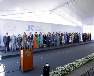 A imagem mostra uma fila de diversas autoridades sobre um palco, durante a cerimônia de entrega da Medalha Presidente Juscelino Kubitschek, em Diamantina. Algumas autoridades vestem roupas formais e outras, uniformes militares da PMMG e do CBMMG. Ao fundo, aparece um grande painel com o brasão do Estado de Minas Gerais, o nome da condecoração e um retrato de Juscelino Kubitschek.