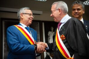 A imagem mostra dois homens se cumprimentando com um aperto de mãos. À esquerda, está o desembargador Osmar Duarte Marcelino, vestindo terno azul, camisa branca, gravata azul e uma faixa cerimonial sobre o corpo. À direita, está o desembargador Sebastião Geraldo de Oliveira, usando toga preta e também uma faixa cerimonial.
