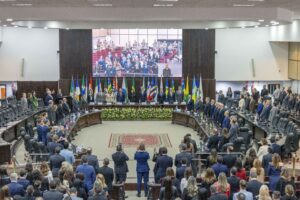 A imagem mostra o Plenário do TJPR, com autoridades e representantes de diferentes países reunidos em torno de mesas dispostas em formato semicircular. Ao fundo, há diversas bandeiras, um telão exibindo participantes do evento e uma mesa de autoridades em pé.