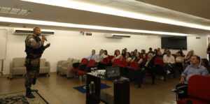 A imagem mostra o auditório do TJMMG com diversas pessoas, entre homens e mulheres, sentadas em fileiras de poltronas vermelhas. À esquerda do auditório está o sargento Kerley Tadeu da Silva Jerônimo, em pé. Ele usa uma farda da Polícia Militar, enquanto fala ao microfone. O auditório tem teto e paredes brancas.