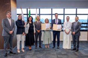 A imagem mostra um grupo de pessoas em pé no Gabinete da Presidência do TJMG, um ambiente formal com parede lateral revestida de madeira, piso escuro e janelas ao fundo, tendo à frente três bandeiras hasteadas. O grupo é formado por oito autoridades dispostas em pé, lado a lado, sendo homens e mulheres que vestem trajes formais. Da direita para a esquerda está o juiz federal Celso Vieira de Souza, vestindo terno cinza, camisa azul clara e gravata cinza; a juíza de Direito Daniela de Freitas Marques, vestida com blusa e calça brancas; o desembargador Fernando José Armando Ribeiro, usando terno azul escuro, camisa clara e gravata roxa; a desembargadora Maria Luiza Santana Assunção, usando vestido cinza claro; o desembargador Vicente de Oliveira Silva, vestindo terno cinza, camisa branca e gravata marrom; a desembargadora Tânia Regina Silva Reckziegel, usando vestido longo preto; a assessora Jéssica Amanda Fachin, usando blusa e blazer escuros e calça branca; e o desembargador André Leite Praça, que está vestindo terno cinza, camisa branca e gravata rosa claro.