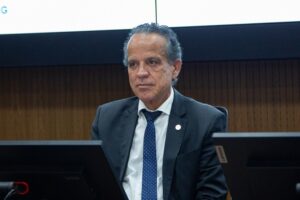 A imagem mostra o desembargador Jâmes Ferreira Santos, vestindo terno cinza-escuro, camisa clara e gravata azul-escuro. Ele está sentado à mesa, diante de duas telas de computador. Ao fundo, aparece uma parede revestida de madeira.
