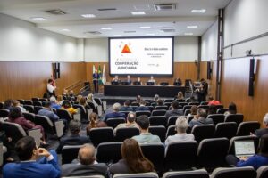 A imagem mostra um auditório da Escola Judicial do TRT-MG com diversas pessoas sentadas, no 2º Encontro de Cooperação Judiciária. Ao fundo, aparece uma mesa de autoridades, no palco, diante de uma tela de projeção, com informações sobre o evento.