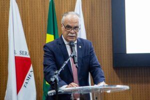 A imagem mostra o desembargador Rúbio Paulino Coelho discursando ao microfone em uma tribuna transparente. Ele está vestindo um terno azul-escuro, uma camisa clara e gravata rosa, e usando óculos. Ao fundo, aparece uma parede revestida de madeira, além de três bandeiras.