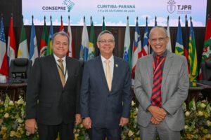 A imagem mostra três homens, em pé, no Plenário do TJPR. O primeiro é o coronel Giovanne Gomes da Silva, vestindo terno preto, camisa branca e gravata bronze; o segundo é o desembargador Osmar Duarte Marcelino, vestindo terno preto, camisa branca e gravata amarela; e o último é o coronel Francisco Valdinei Duarte, que está vestindo terno cinza-claro, camisa cinza-escuro e gravata listrada nas cores preta e vermelha. Ao fundo, aparece parte de um telão exibindo a identidade visual do XXII Encontro do Consepre, além de diversas bandeiras dispostas lado a lado e uma mesa com arranjos de flores.