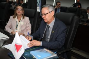 A imagem mostra o desembargador Osmar Duarte Marcelino sentado em uma cadeira, assinando a Carta de Curitiba com uma caneta azul. Ele está vestindo um terno escuro, camisa cinza-clara e gravata escura. À sua frente, aparecem documentos e uma pasta azul sobre a mesa, além de uma mini bandeira de Minas Gerais.