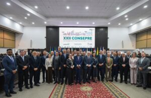 A imagem mostra um grupo de diversas pessoas, entre homens e mulheres, em pé, no TJPR. O ambiente tem paredes brancas e um painel de madeira ao fundo. Sobre o painel, há um telão exibindo o título e a identidade visual do XXII Encontro do Consepre.