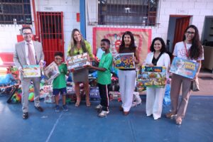 A imagem mostra sete pessoas, sendo duas crianças, segurando caixas de brinquedos, no pátio da Escola Municipal Tristão da Cunha. Da direita para a esquerda, a segunda pessoa é a juíza de Direito Renata Rodrigues de Pádua, vestindo blusa preta, um casaco branco e calça branca, e segurando uma caixa de brinquedo. Ao fundo, aparecem diversas caixas de brinquedos no chão. O pátio da escola possui parede branca e piso azul. 
