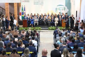 A imagem mostra um palco com diversas pessoas sentadas em cadeiras, todas vestidas com roupas formais. Ao fundo, aparecem três bandeiras e um painel com a identidade visual do evento. Nele está escrito: Governo de Minas. Medalha Santos Dumon. Em primeiro plano, aparecem diversas pessoas sentadas em fileiras de cadeiras, voltadas para o palco.