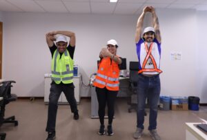 A imagem mostra três pessoas em pé, em uma sala do TJMMG, realizando exercícios de alongamento. À esquerda, um homem alonga o braço, puxando o cotovelo por trás da cabeça, com uma perna posicionada à frente e a outra estendida para trás. Ao centro, uma mulher mantém um braço estendido à frente enquanto o puxa uma mão em direção ao corpo com a outra mão. À direita, aparece um homem com os braços esticados acima da cabeça. O ambiente possui parede branca e piso bege.